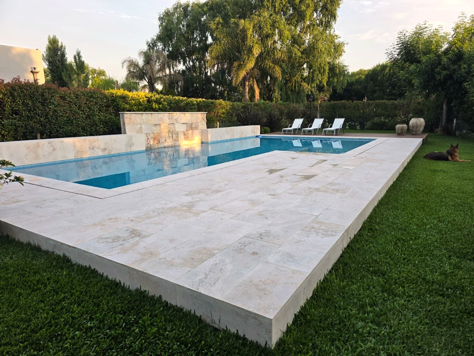 Pileta con borde y solado de travertino sobre el pasto, al atardecer