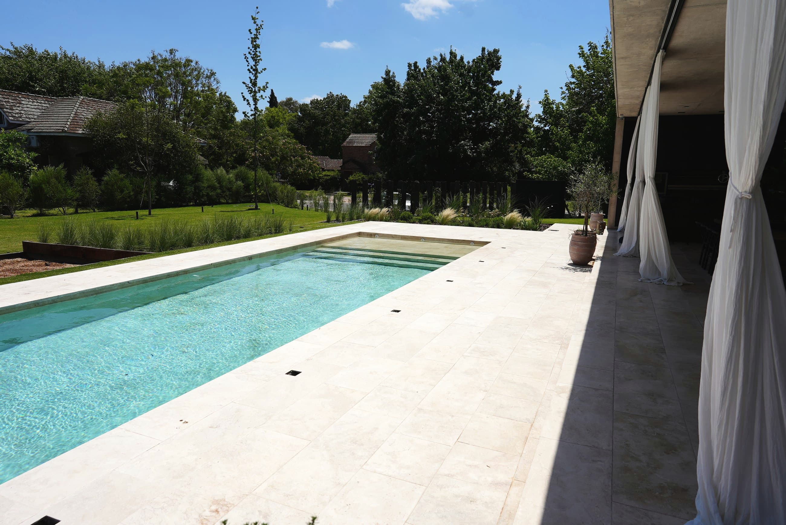 Pileta con solado de travertino junto a una galería, a pleno sol