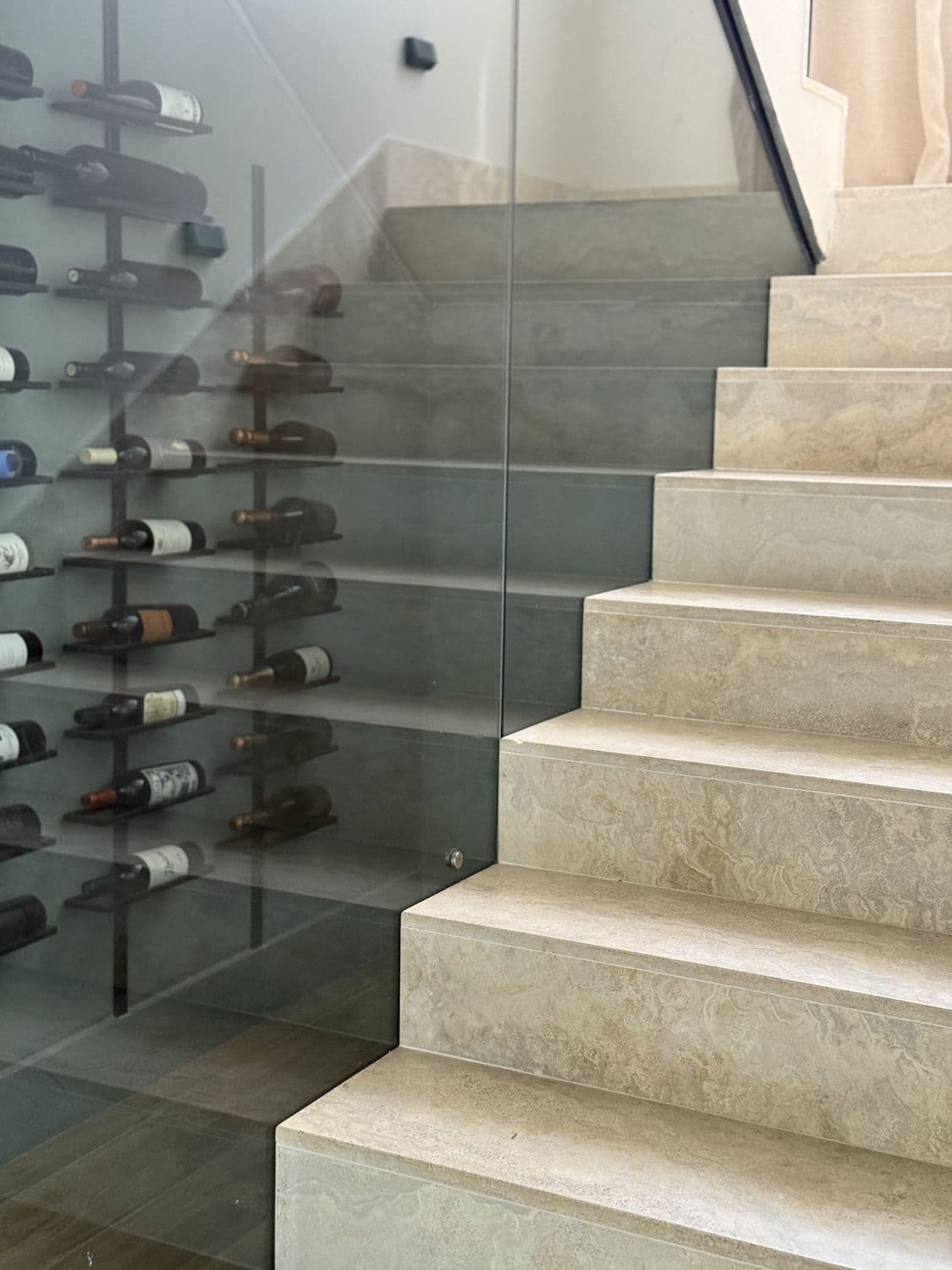 Escalera maciza de travertino con baranda de vidrio y la cava de vinos detrás